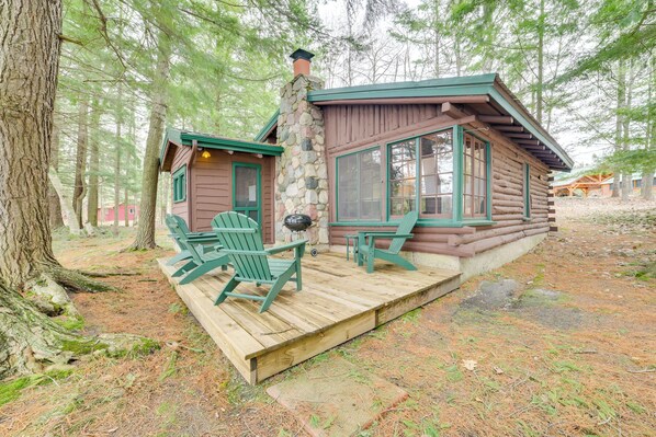 Una cabaña de troncos con una terraza de madera, dos sillas Adirondack verdes y una chimenea de piedra.