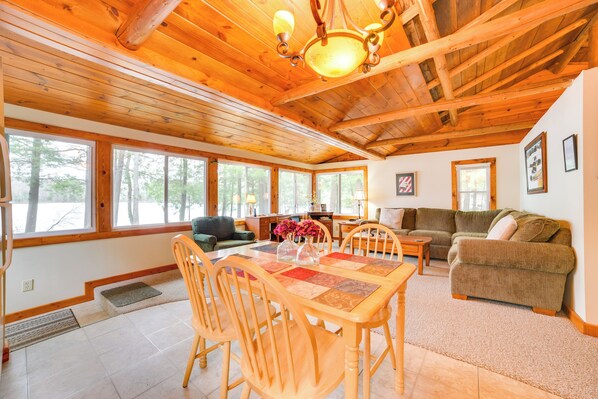 A spacious living area with a wooden ceiling, large windows, a dining table with chairs, and a sofa.
