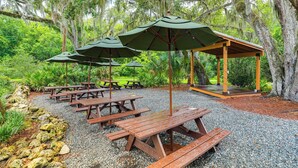 A covered picnic area with wooden benches and green umbrellas, surrounded by lush greenery and trees. - Walk to Community Perks: Home in The Villages (The Villages)