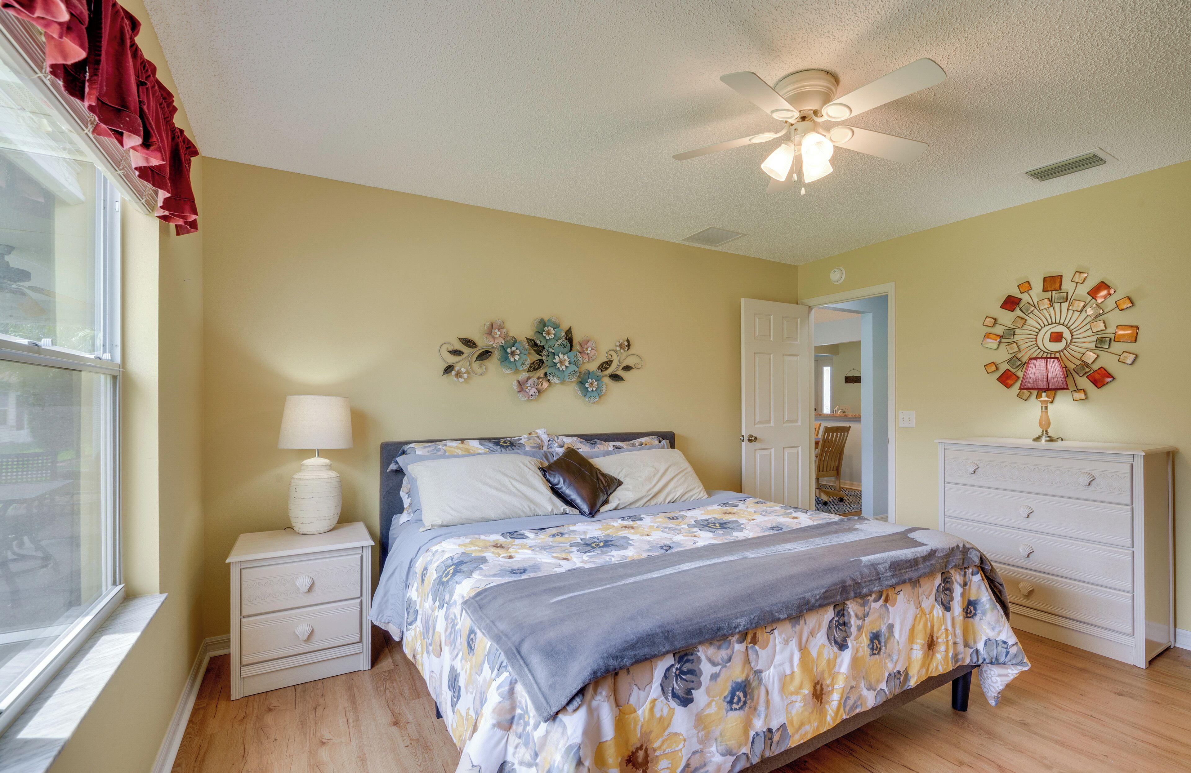 Une chambre à coucher comprenant un lit, des tables de chevet, une lampe, un ventilateur de plafond, une fenêtre avec des rideaux et un élément décoratif mural.