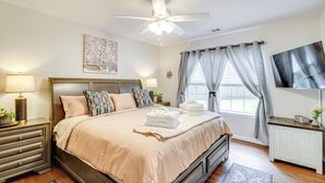 A neatly arranged bedroom with a bed, bedside lamps, a ceiling fan, a TV, and a window with curtains. - Deck & Yard: Savannah Area Retreat! (Pooler)