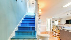 A staircase with blue steps leading to a door, a white chair, a sofa, and a lamp in a living room area. - Hot Tub & Bay Views: Outer Banks Home (Kill Devil Hills)