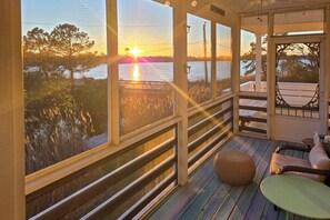 Een overdekt terras met uitzicht op een zonsondergang boven het water, houten leuningen en buitenmeubilair.