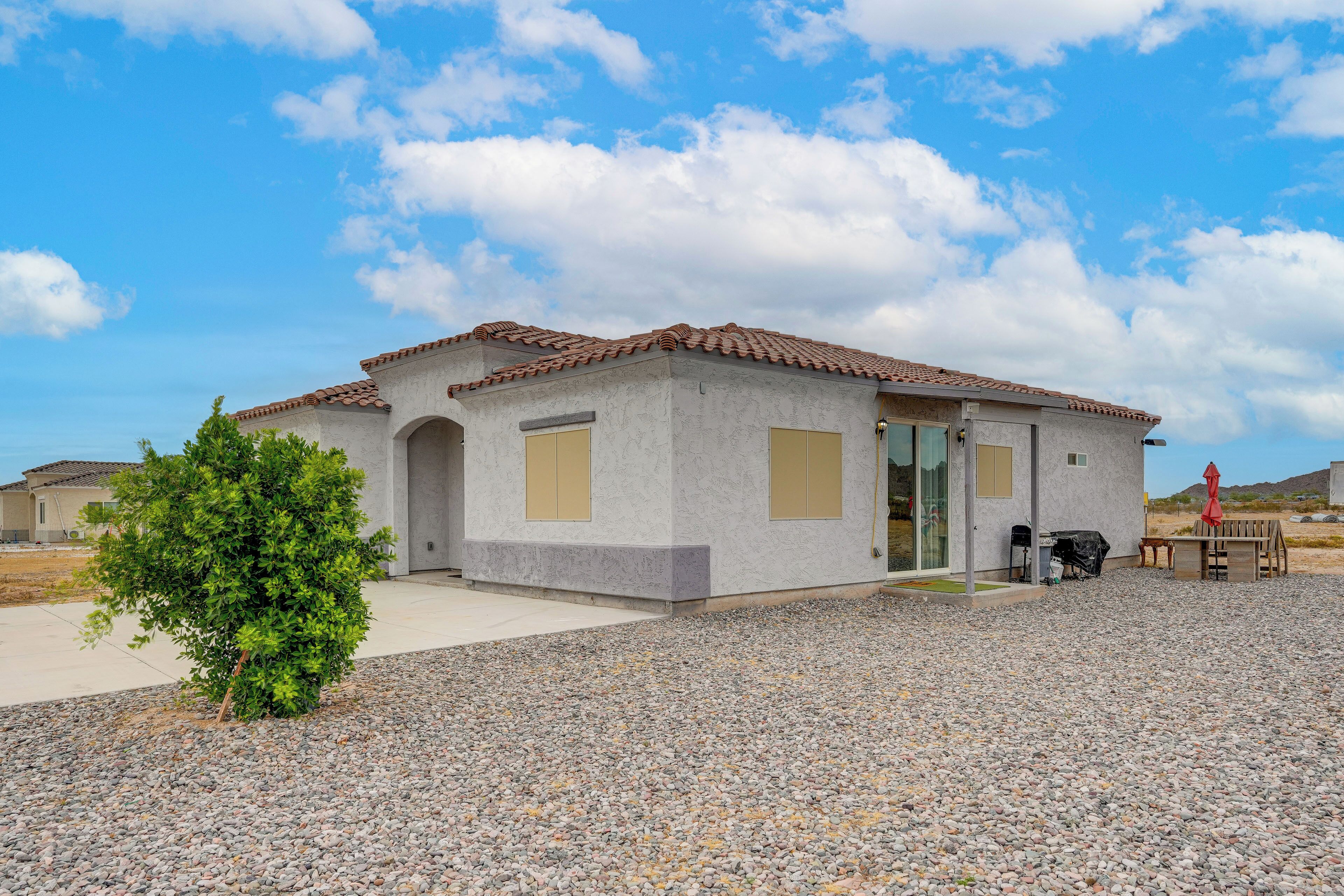 A single-story house with a tiled roof, a small patio area with a table and chairs, and a gravel driveway.