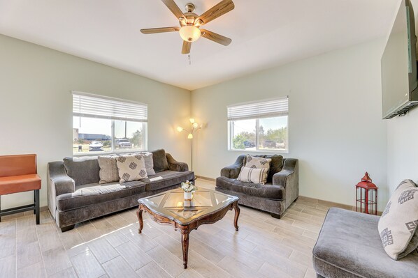 A spacious living room with two grey sofas, a glass coffee table, a wooden ceiling fan, and large windows with blinds. - Mtn Views: Spacious Family Home in Casa Grande! (Casa Grande)