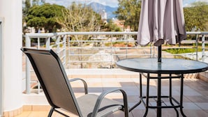 Ein Balkon mit Stuhl und Tisch, ein grauer Sonnenschirm und Blick auf Bäume und ein Berg.