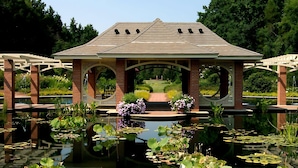 A gazebo with a brick base and a tiled roof, surrounded by a pond with lily pads and flowers.