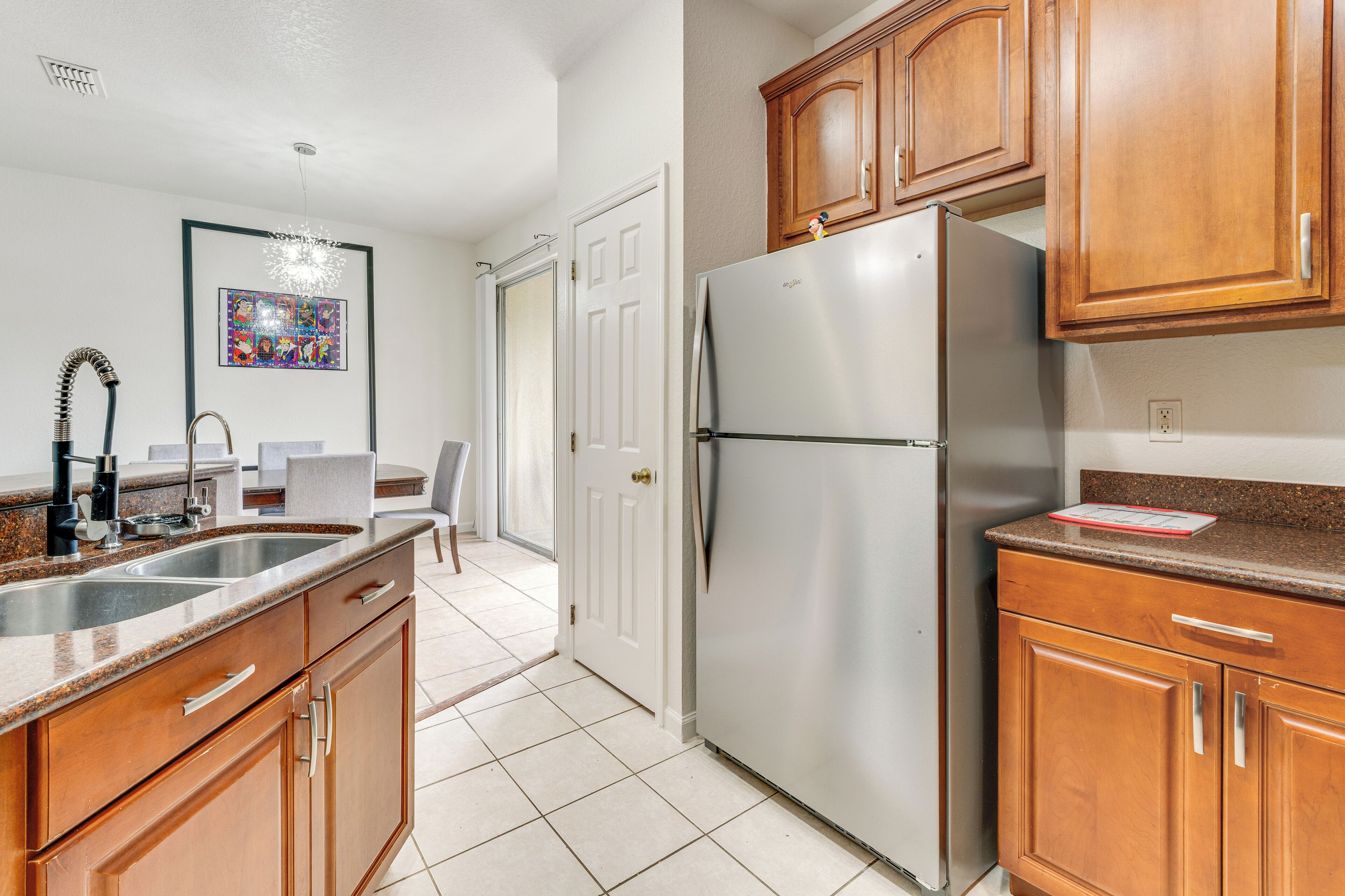 A kitchen with wooden cabinets, a stainless steel refrigerator, a double sink, and a framed artwork on the wall.