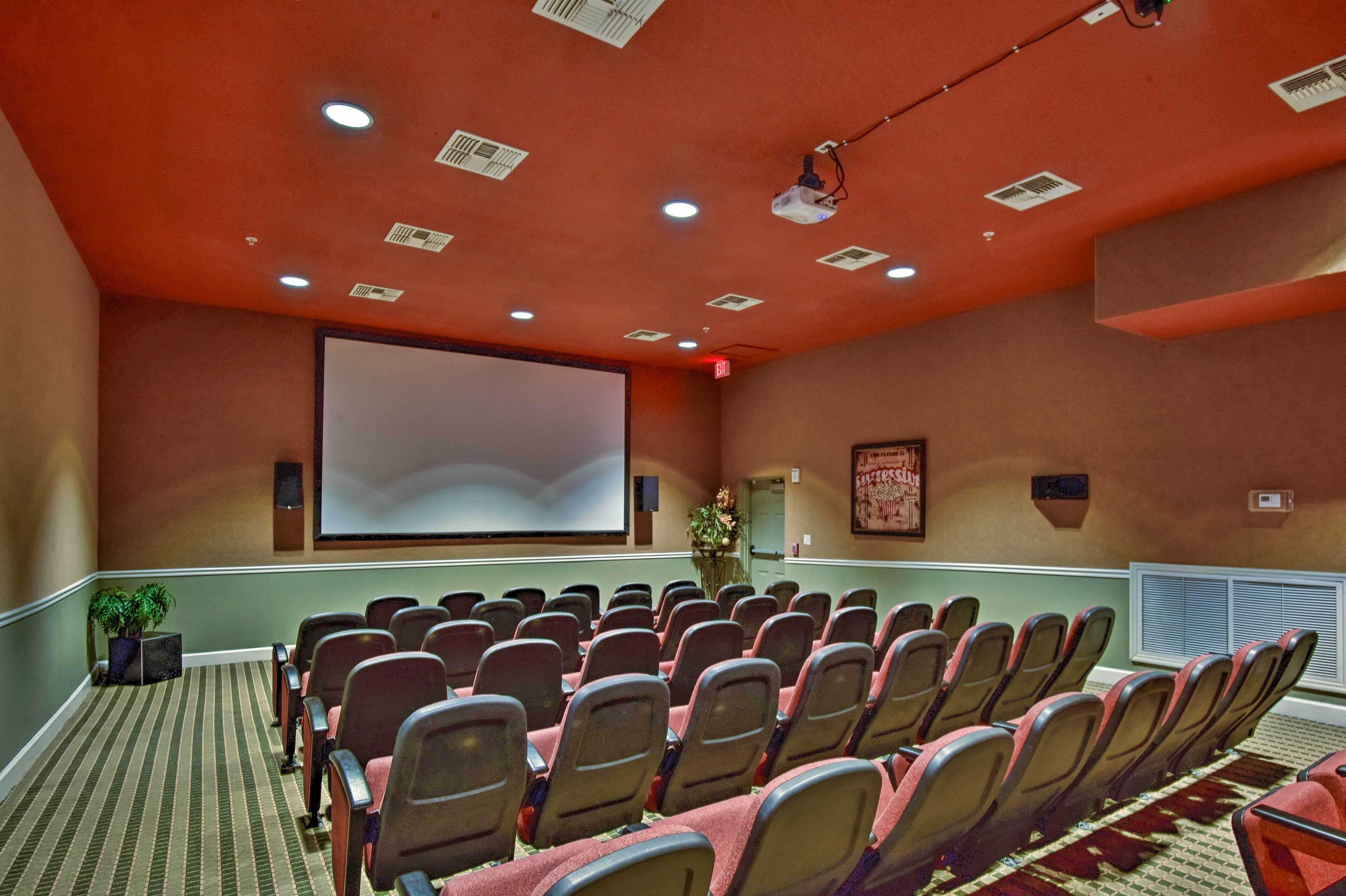 Un salón de conferencias con filas de sillas, una pantalla de proyector y un ventanal con persianas.