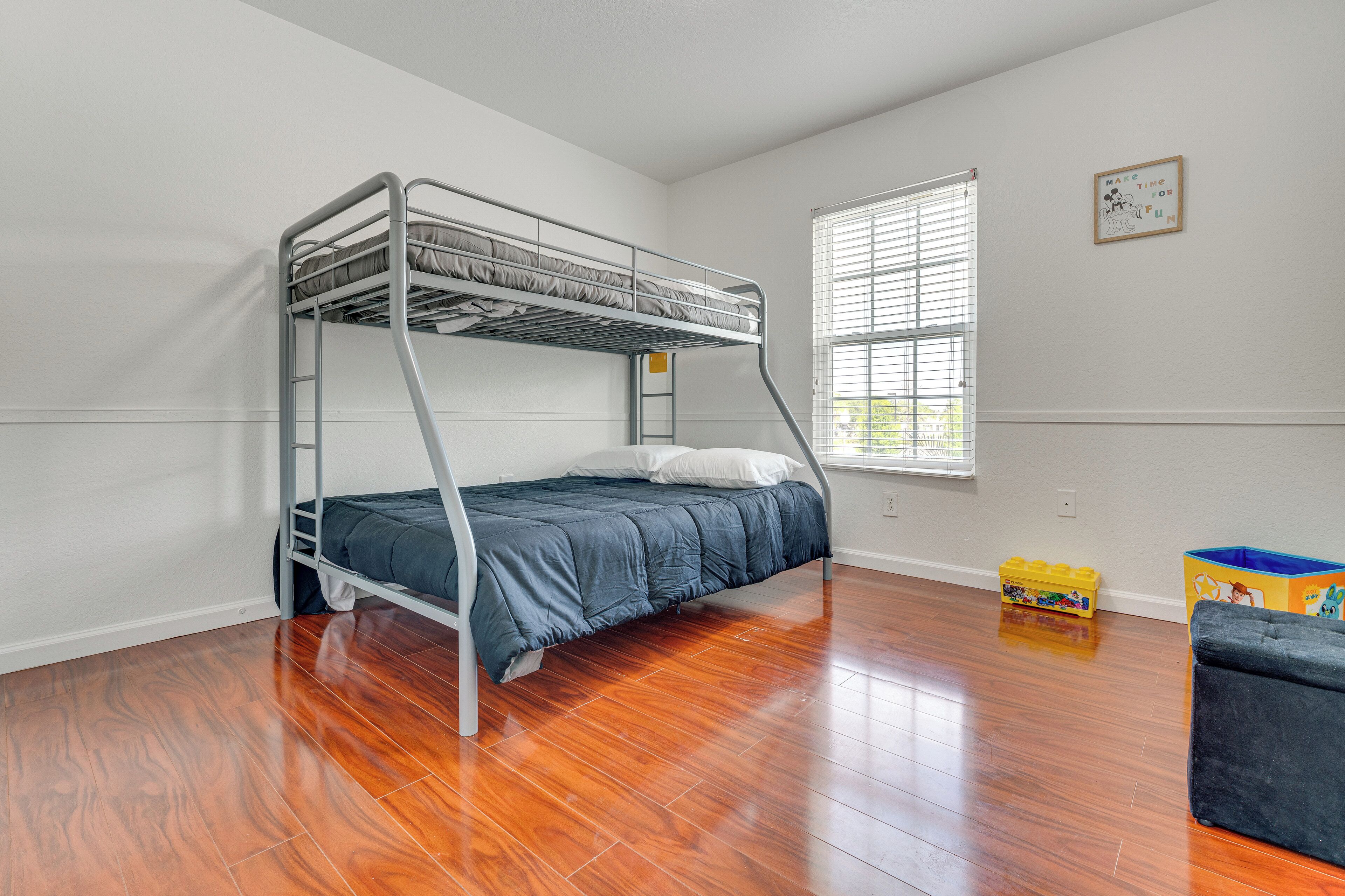 Un lit superposé dans une chambre avec un plancher en bois, une fenêtre avec des stores et un espace de jeux pour enfants avec des jouets.