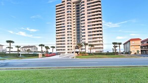 A tall, multi-story building with a modern design, featuring a prominent entrance and a well-maintained surrounding area with grass and palm trees.