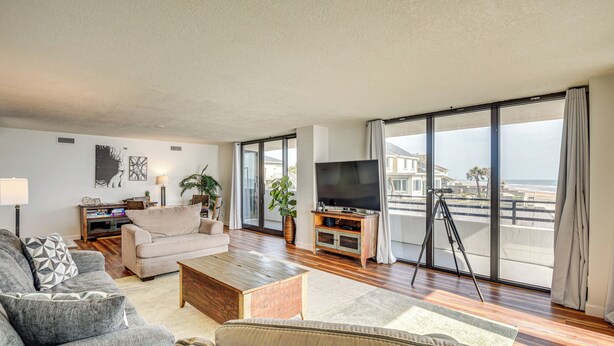 A spacious living room with a large TV, comfortable seating, a wooden coffee table, and a balcony with a view of the beach.