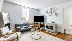 A living room with a sofa, armchair, TV, fireplace, and shelves with decorative items. - Explore Dtwn, Parks & Colleges: Cozy Worcester Apt (Worcester)