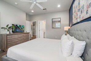 A bedroom with a bed, a television on a dresser, a ceiling fan, and a door leading to another room.