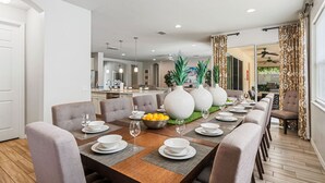 A modern dining area with a long table set for six, surrounded by comfortable chairs. There are large white vases on the table, and a bowl of fruit. The room has a clean, minimalist design with white walls and a wooden floor.