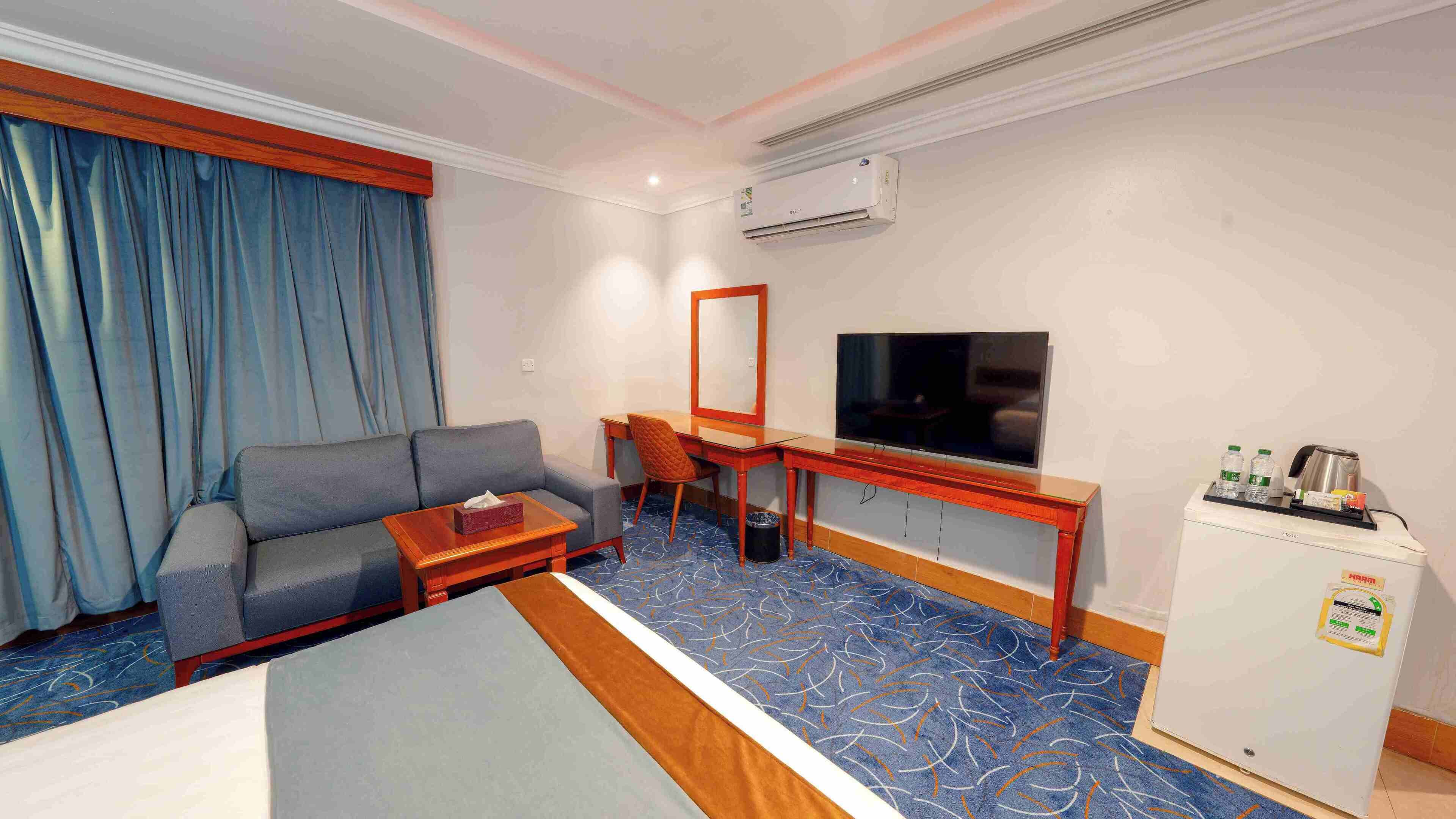A hotel room with a bed, a sofa, a desk with a chair, a television, and a minibar.