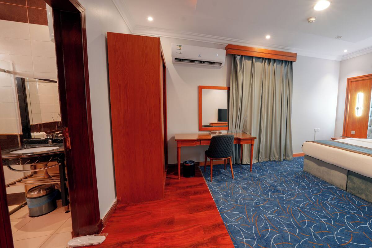 A hotel room with a bed, a desk, a chair, a bathroom, and a red carpet.