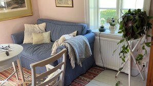 Een gezellige kamer met een geruite bank, een witte stoel, een klein tafeltje, een tapijt met dessin en een raam met uitzicht op groen.