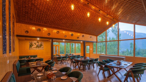A restaurant with wooden walls, green seating, and large windows offering a view of the mountains. - Moustache Pahalgam (Anantnag)