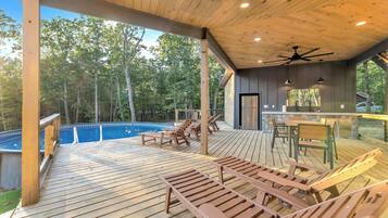 A covered outdoor deck with wooden furniture, a pool, and a stone wall.