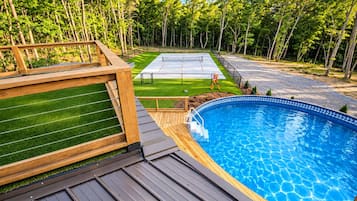 A swimming pool with a wooden deck and a grassy area with a tennis court.