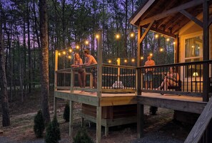Eine Holzterrasse mit Lichterketten, einer überdachten Sitzecke und einem kleinen Tisch, mit Blick auf den Wald.
