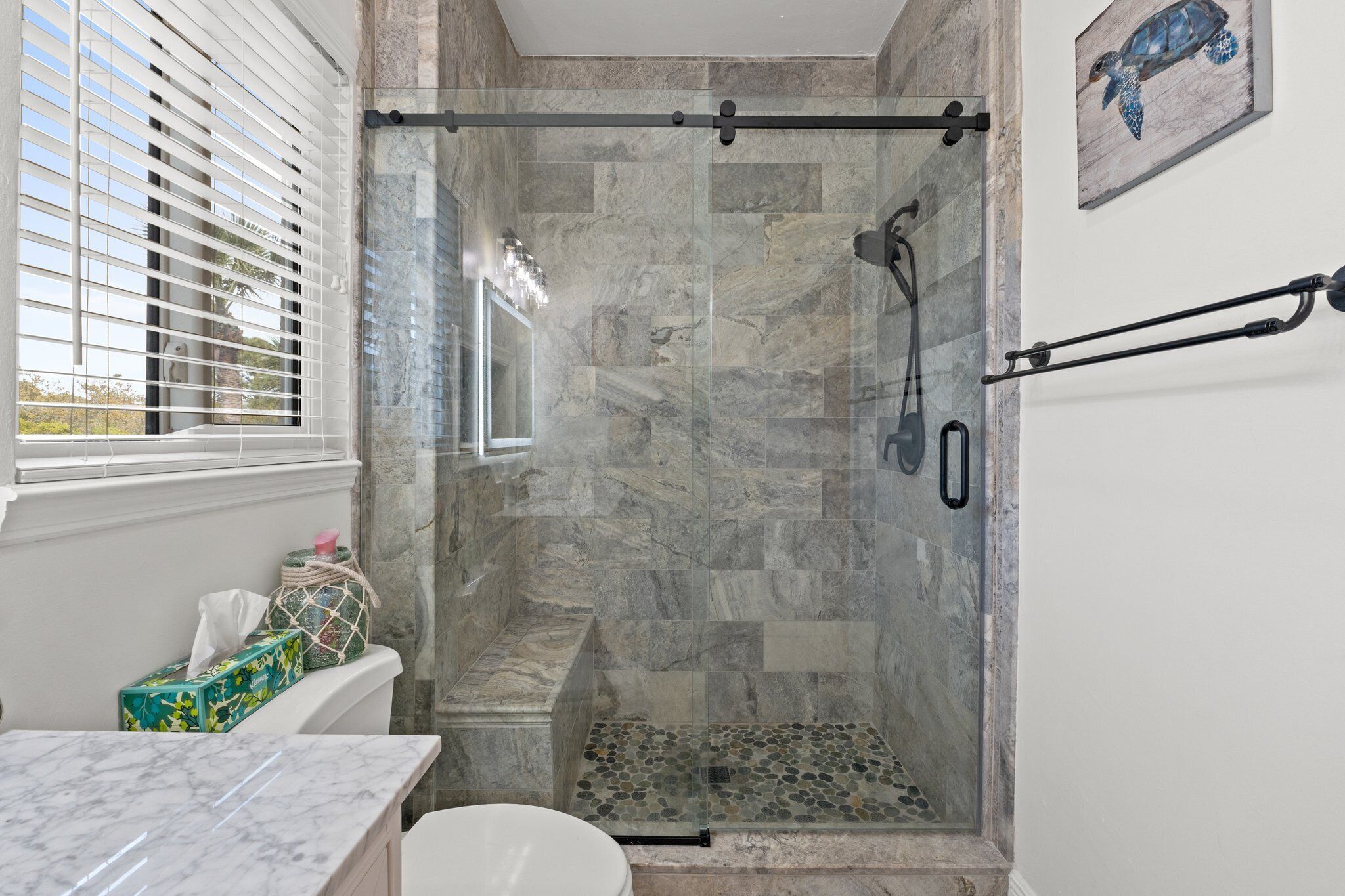 Un baño moderno con ducha de vidrio, paredes de mármol y ventana con persianas.
