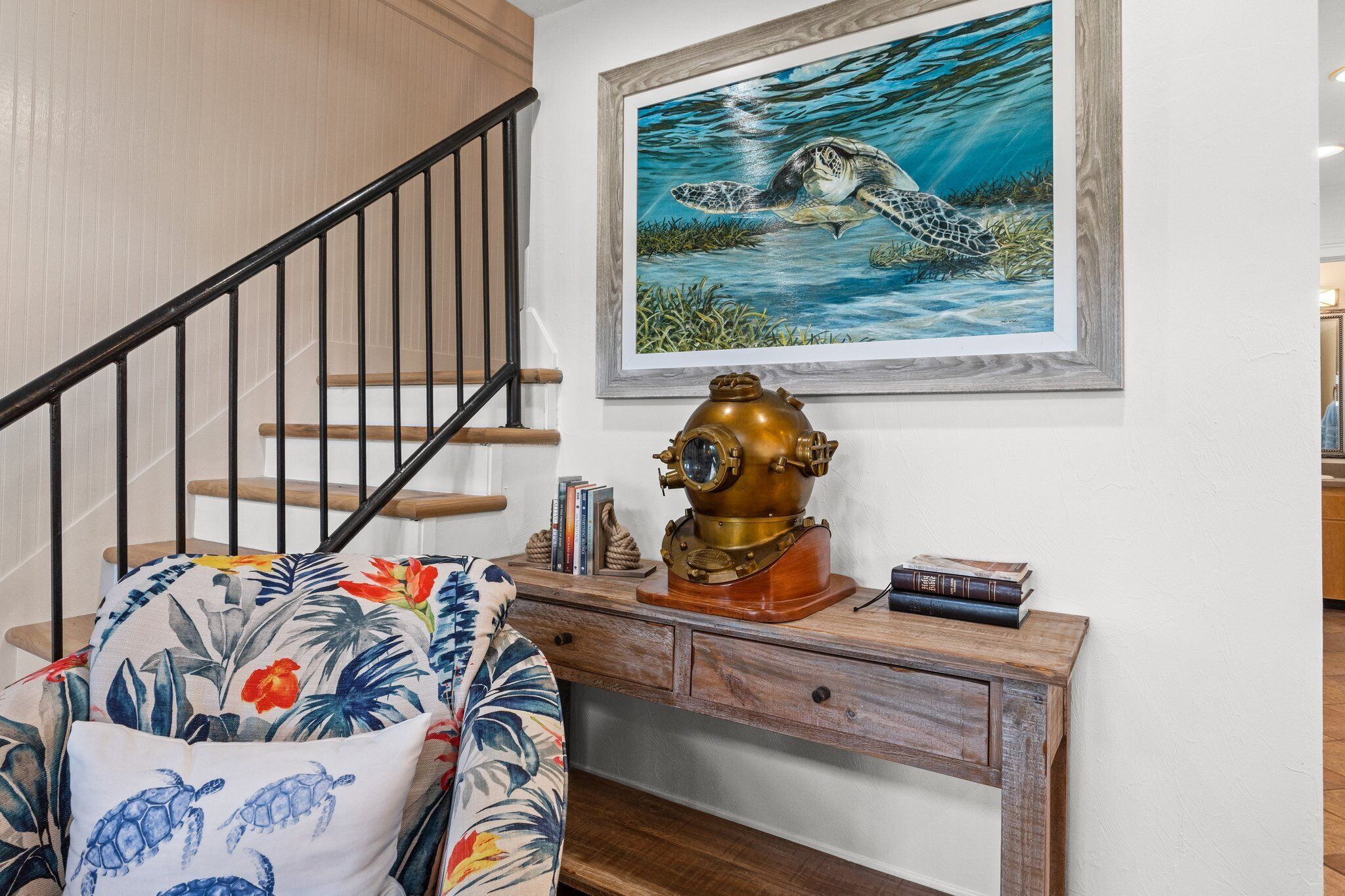 A staircase with a wooden handrail, a wall-mounted painting of a sea turtle, and a wooden desk with a brass object and books.