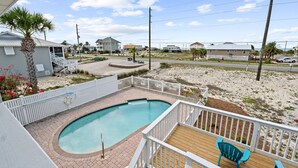 A swimming pool with a wooden deck and white railings, surrounded by a sandy area and a few buildings in the background.