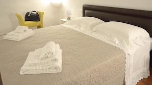 Ein ordentlich bezogenes Bett mit weißen Leinen, ein gelber Sessel und ein kleiner Nachttisch mit Lampe.