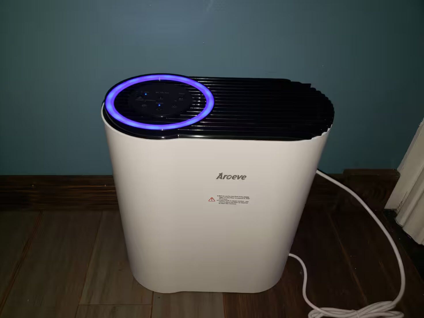 A white portable air purifier with a blue light on top, labeled "Aroeve," placed on a wooden floor against a teal wall.