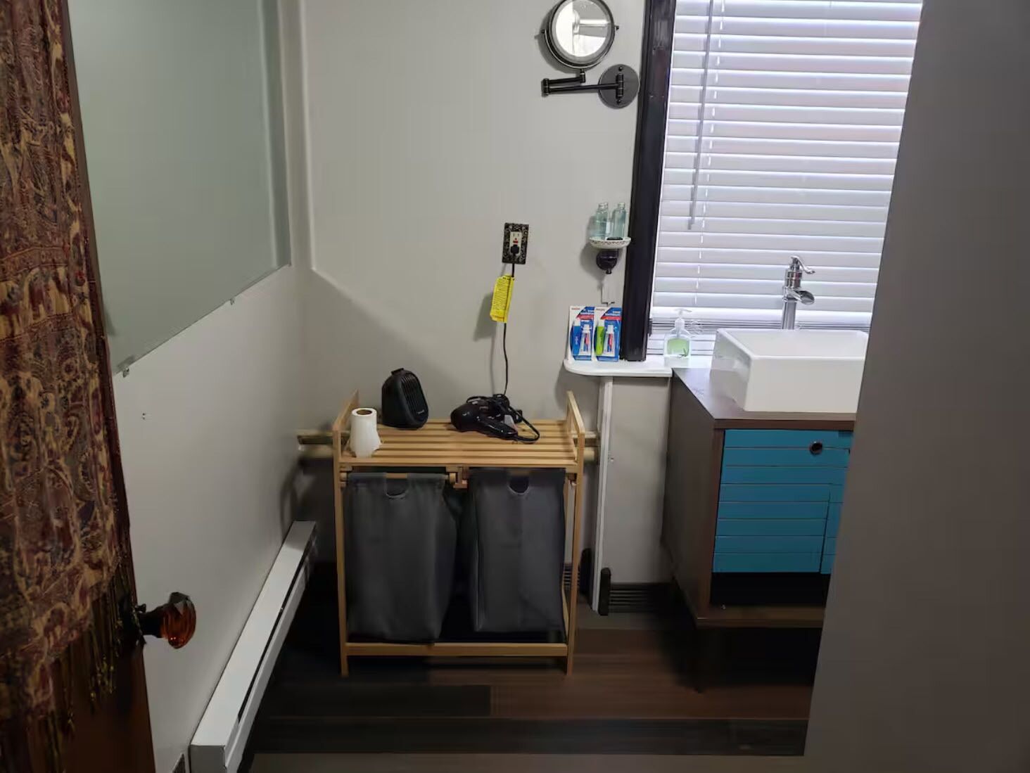 Un baño pequeño con un tendedero de madera, un lavamanos con espejo y una ventana con persianas.