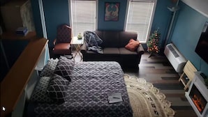 A compact bedroom with a bed, a sofa, a small table, a chair, a ceiling fan, a fireplace, and a Christmas tree. - Muskegon Lake View Loft studio/kitchenette (Muskegon)