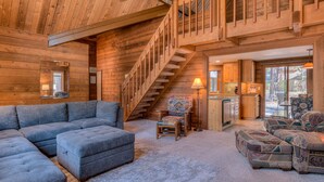 A log cabin interior with a staircase, a living room with a sectional sofa, a patterned armchair, and a kitchen area.