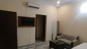 A hotel room with a bed, a sofa, a coffee table, a TV mounted on the wall, and an air conditioning unit. - Hotel Crown Plaza (Karachi)