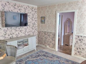 Une pièce avec un papier peint à motifs floraux, un téléviseur fixé au mur, une commode avec des tiroirs, un miroir et un tapis à motifs.
