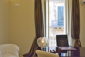 Ein Zimmer mit einem weißen Stuhl, einem Tisch mit einer Lampe und Blick auf einen Balkon mit blauen Fensterläden.