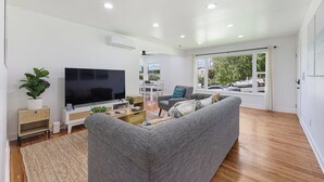 A modern living room with a flat-screen TV, grey sofa, wooden flooring, and large windows. - 4BR | Hilltop House Sunset Cliffs | Beach, Bars, Ocean View (San Diego)
