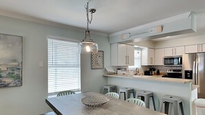 A modern kitchen with white cabinets, a stainless steel refrigerator, and a dining area with a wooden table and bar stools. - Serenity Cottage | Gulfside Community, Private Pool + Bikes (Miramar Beach)