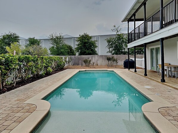 Une piscine rectangulaire, bordée d’un revêtement de carrelage, entourée d’une clôture en bois et de verdure.