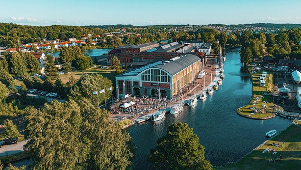 A riverside building with a large glass facade, surrounded by a dock with boats, and a park area with people. - Charming Swedish Forest Cottage Motala (Motala)