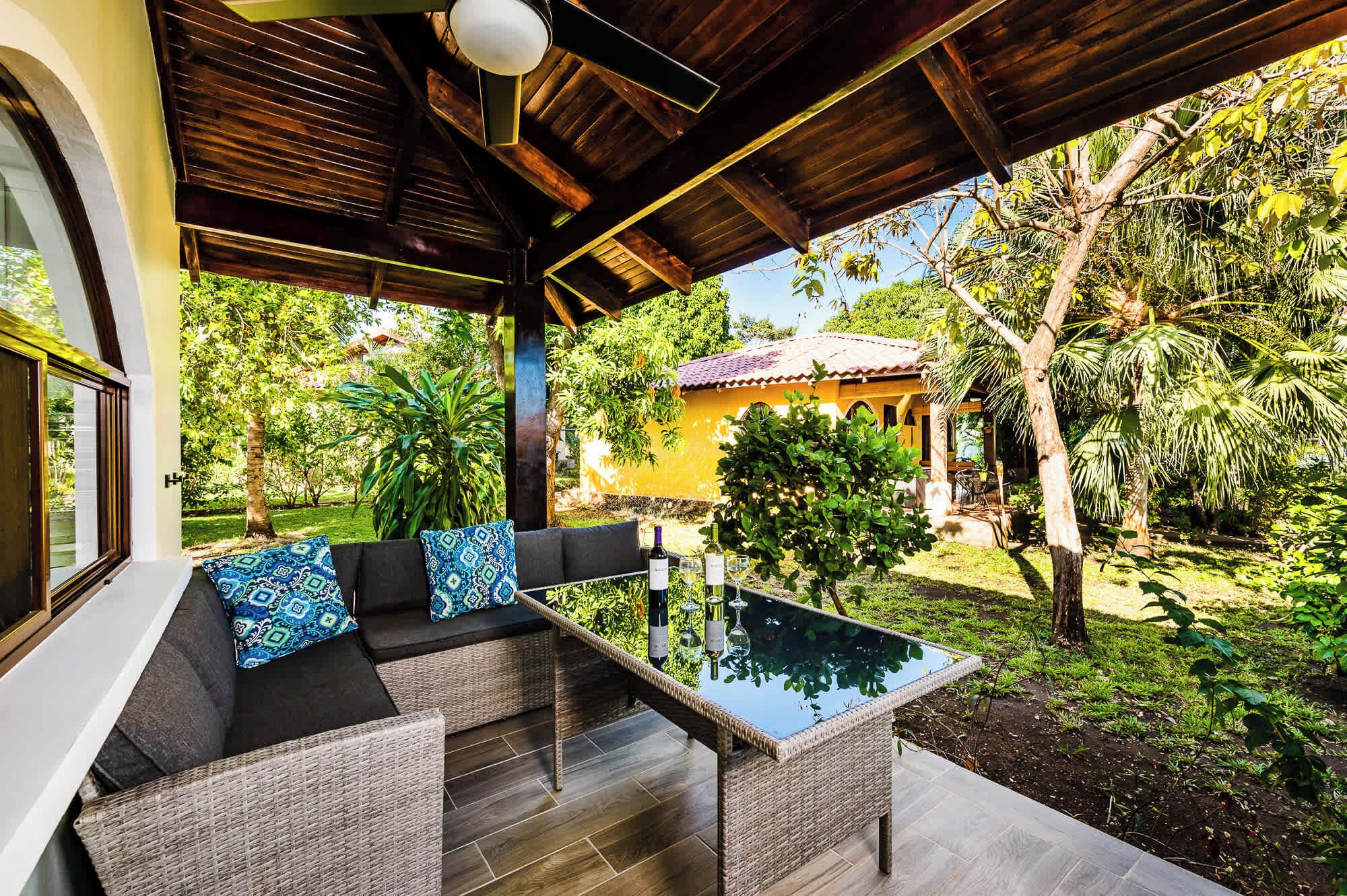 A covered outdoor seating area with a glass-top table, wicker furniture, and a view of a lush garden and a yellow building in the background.