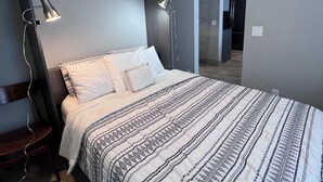 Un dormitorio con cama, lámparas de noche, una silla y una cocina visible al fondo.
