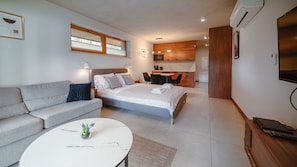 A modern hotel room with a bed, sofa, kitchenette, and a TV.