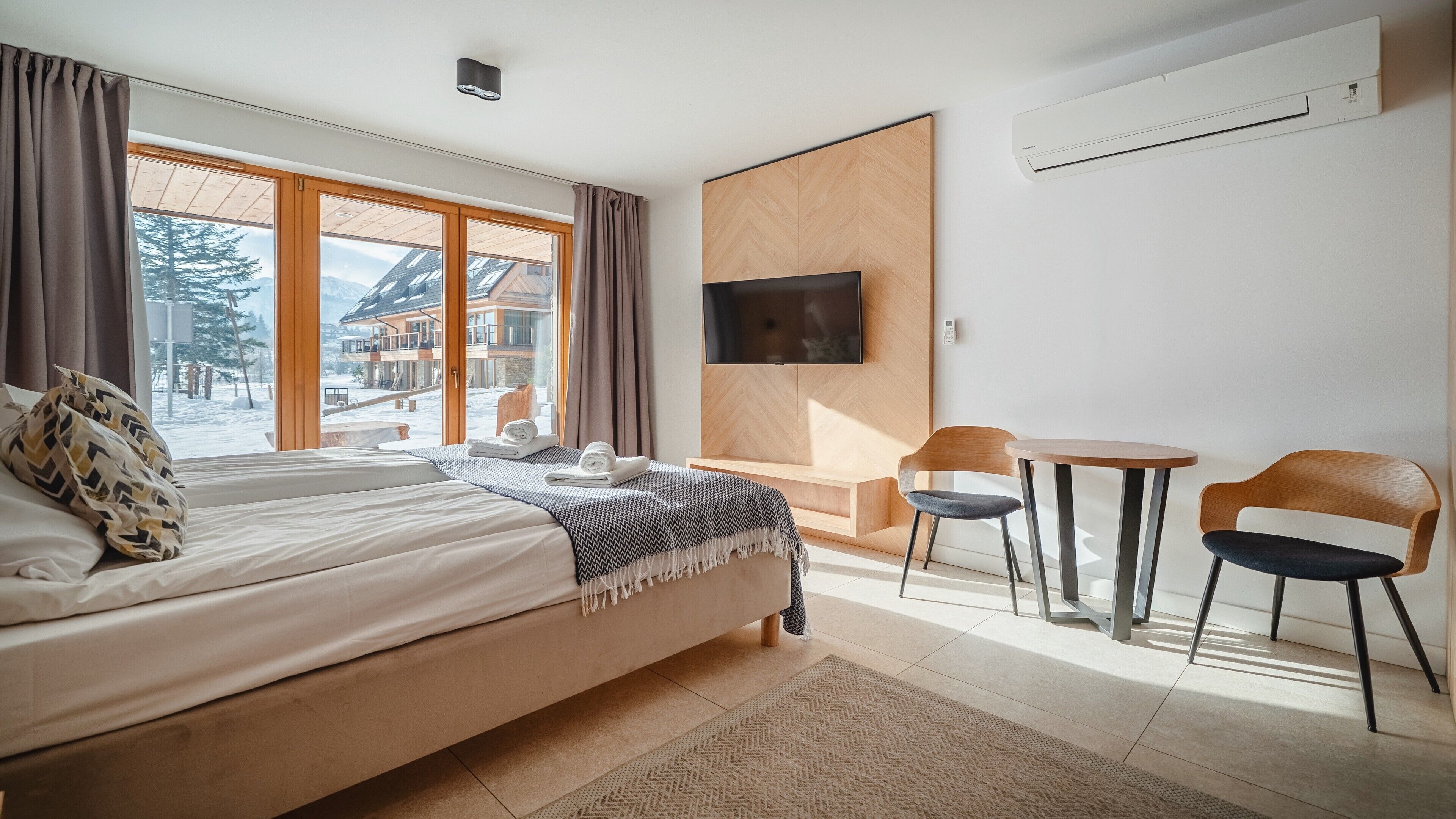 Habitación de hotel con una cama grande, televisión montada en la pared, una mesita con dos sillas y vistas a un paisaje nevado a través de la ventana.