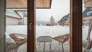 Vista desde una habitación con puerta de madera que da a un área exterior cubierta de nieve, con sillas de mimbre y una pequeña mesa.