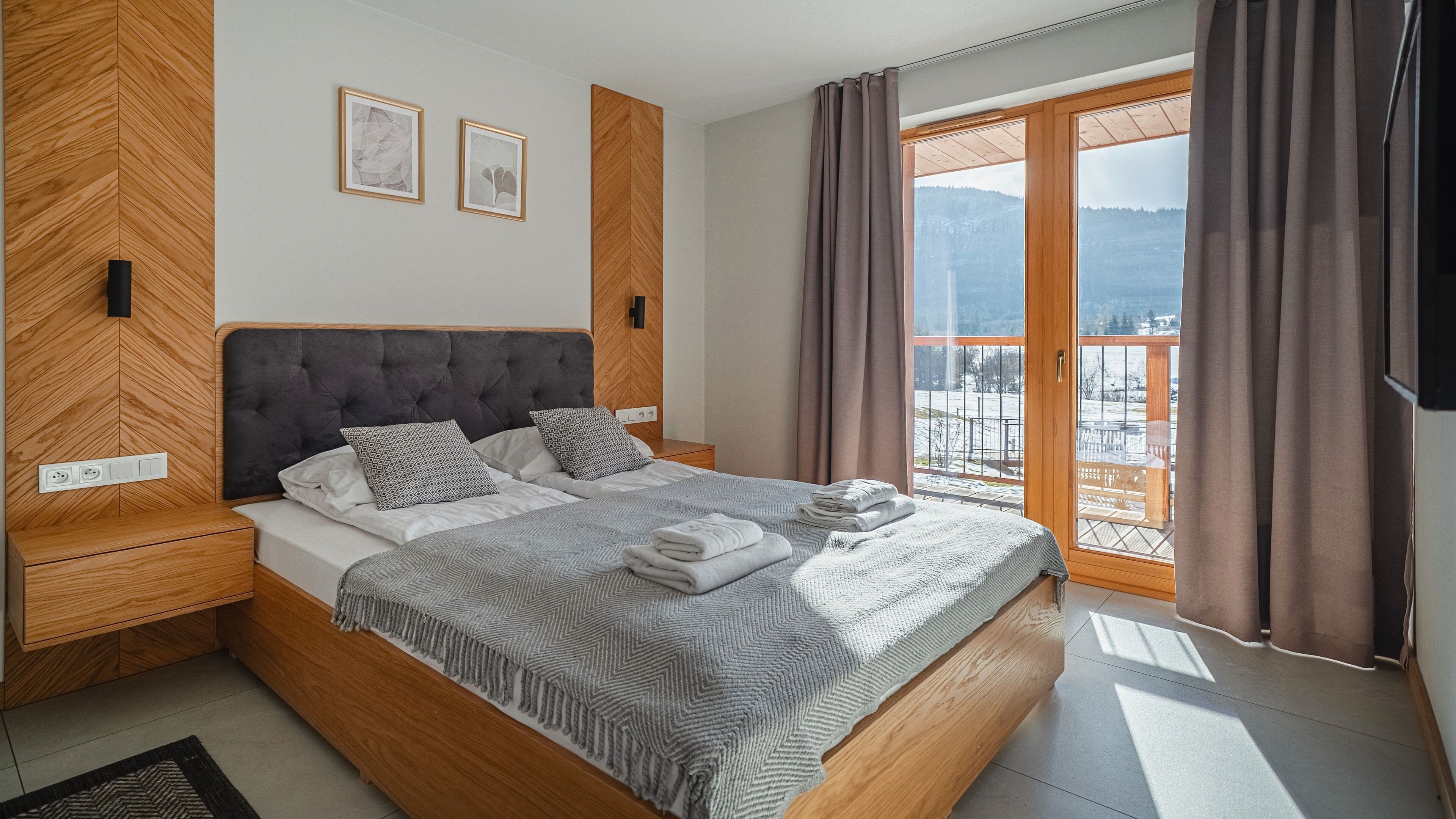 Un dormitorio moderno con una cama grande, cabecera de madera, dos mesitas de noche y vistas a un paisaje nevado a través de un ventanal amplio. La pared presenta un patrón de espiga.