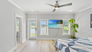 A bedroom with a bed, a ceiling fan, a large window with a view of a beach, and a door leading to another room.