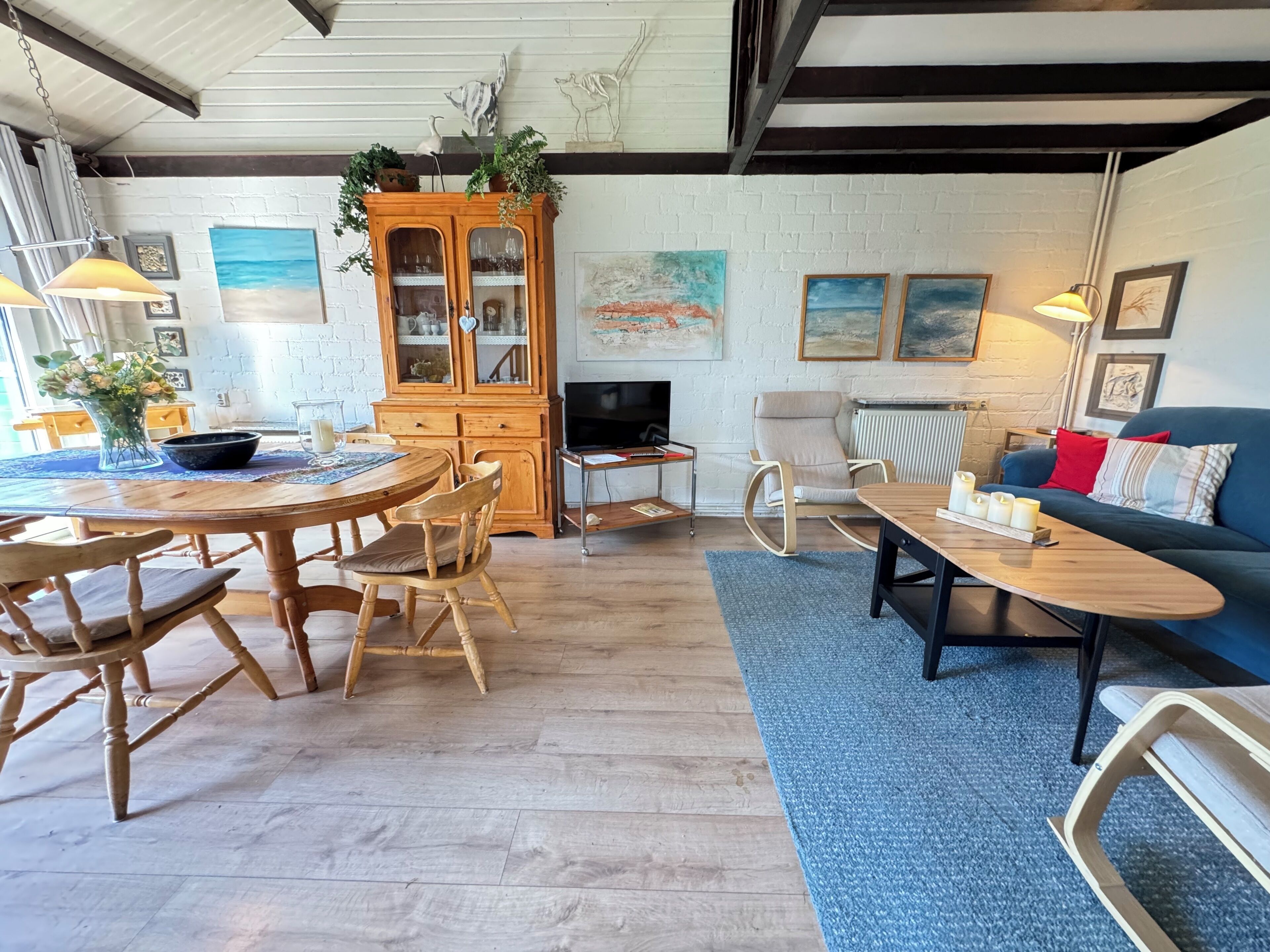 A cozy living room with a wooden dining table, chairs, a blue sofa, and a rocking chair. A cabinet with glass doors and a television are also visible.