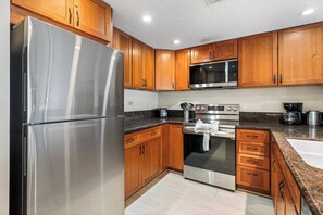 A modern kitchen with stainless steel appliances, wooden cabinets, granite countertops, and a built-in microwave. - Beachfront Bliss at Kona By The Sea #312 - Saltwater Pool & Central AC (Kailua-Kona)